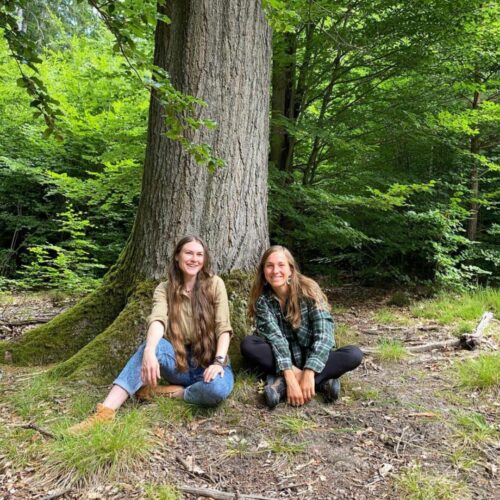 Emely von der Naturakademie Heidelberg und Verena sitzen an einem Baum und lächeln in die Kamera.